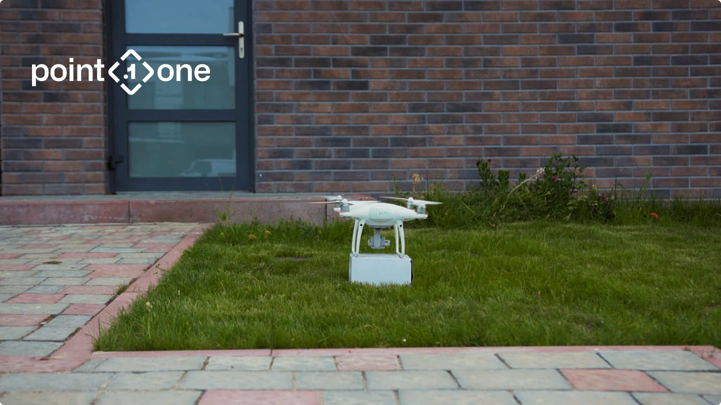 Delivery drone carrying a parcel on a lawn with the Point One Navigation logo, from Point One's drone solutions page